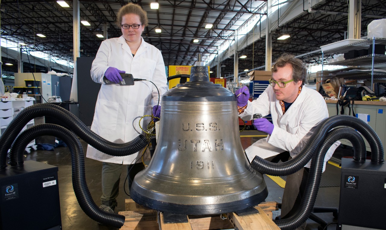 Melissa Swanson and Karl Knauer, conservators at Naval History and Heritage Command, undertake conservation cleaning on the exterior of the USS Utah bell. (U.S. Navy photo by Naval History and Heritage Command Collection Management Facility staff/Released)